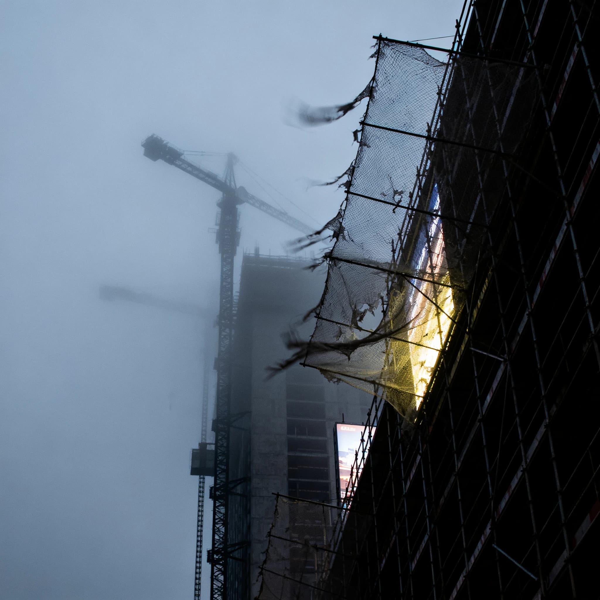 A construction site stretched upward into fog, cranes frozen in silhouette. Wind rattled scaffolding draped with torn mesh that glowed faintly from nearby billboards