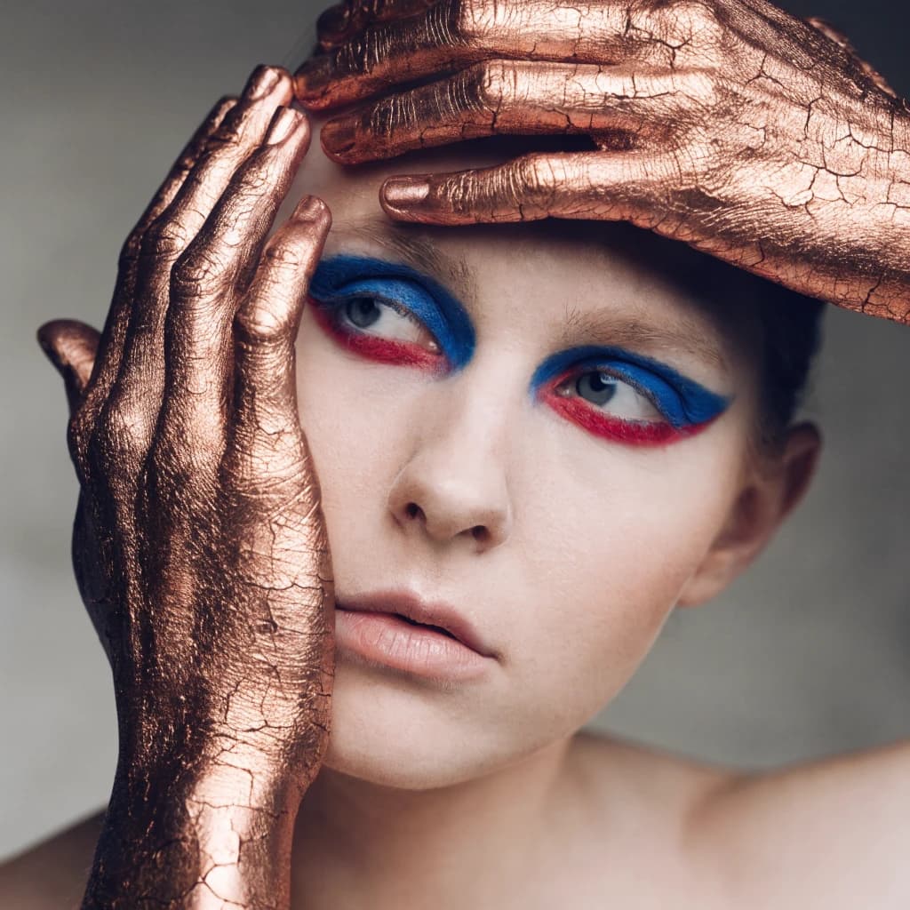 A striking close-up portrait of a woman with cracked metallic copper-painted hands framing her face. Her vivid blue and red eye makeup contrasts sharply with the smooth, pale skin and muted background, creating a bold, surreal composition. She displays an air of uncertainty about her