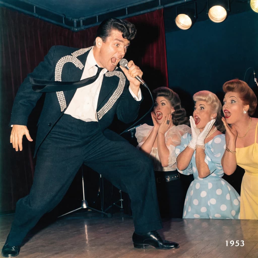 An archival image from 1953 of the rock singer swinging his hips on the stage while glamorous back up singers feign shock at his gyrations! He wears a diamond encrusted jacket with the stones arranged in the shape of a heart