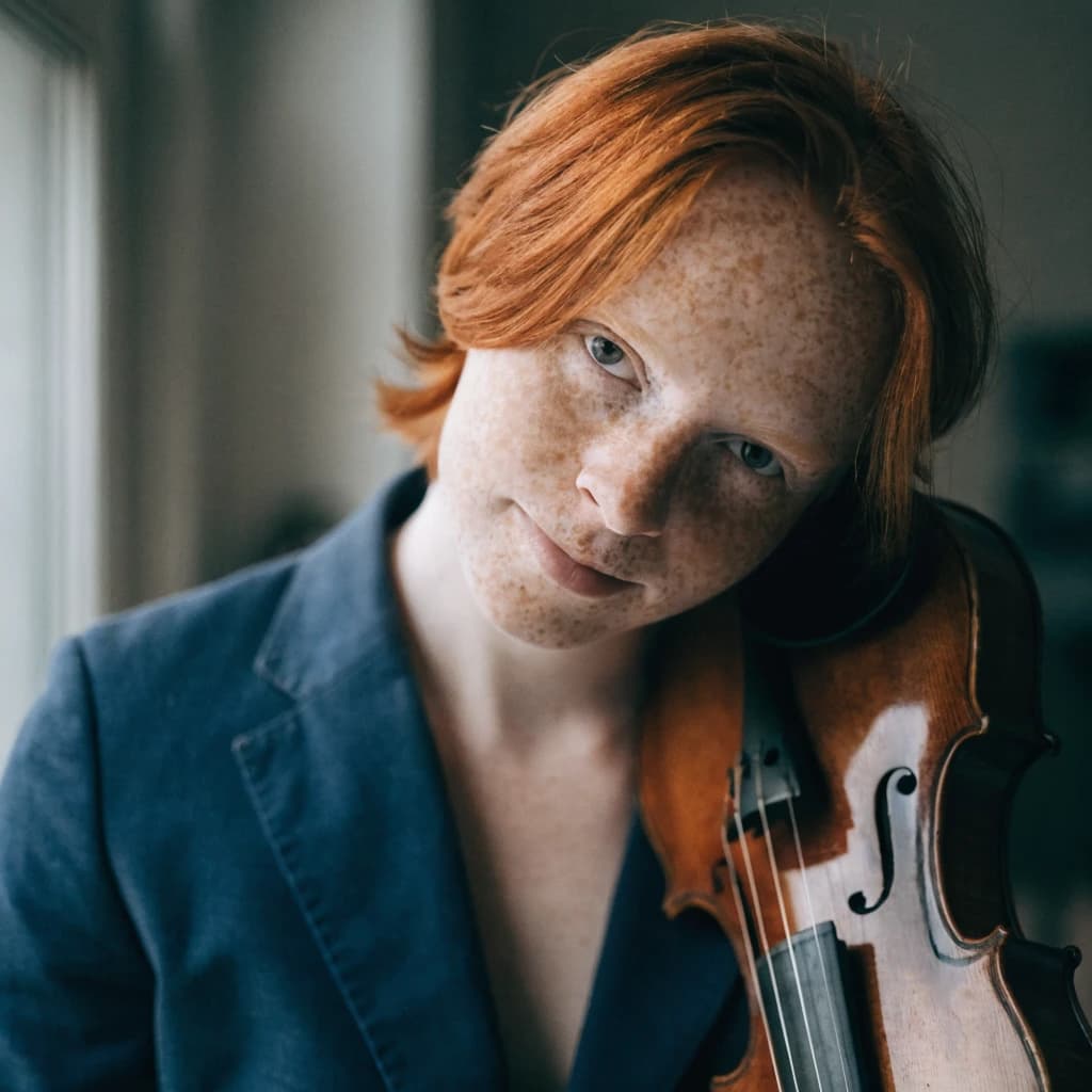 Capture a head-and-shoulders portrait of a freckled red-haired violinist in a navy blazer, soft window light, 85mm at f/1.8, gently smiling yet serious eyes, muted tones.