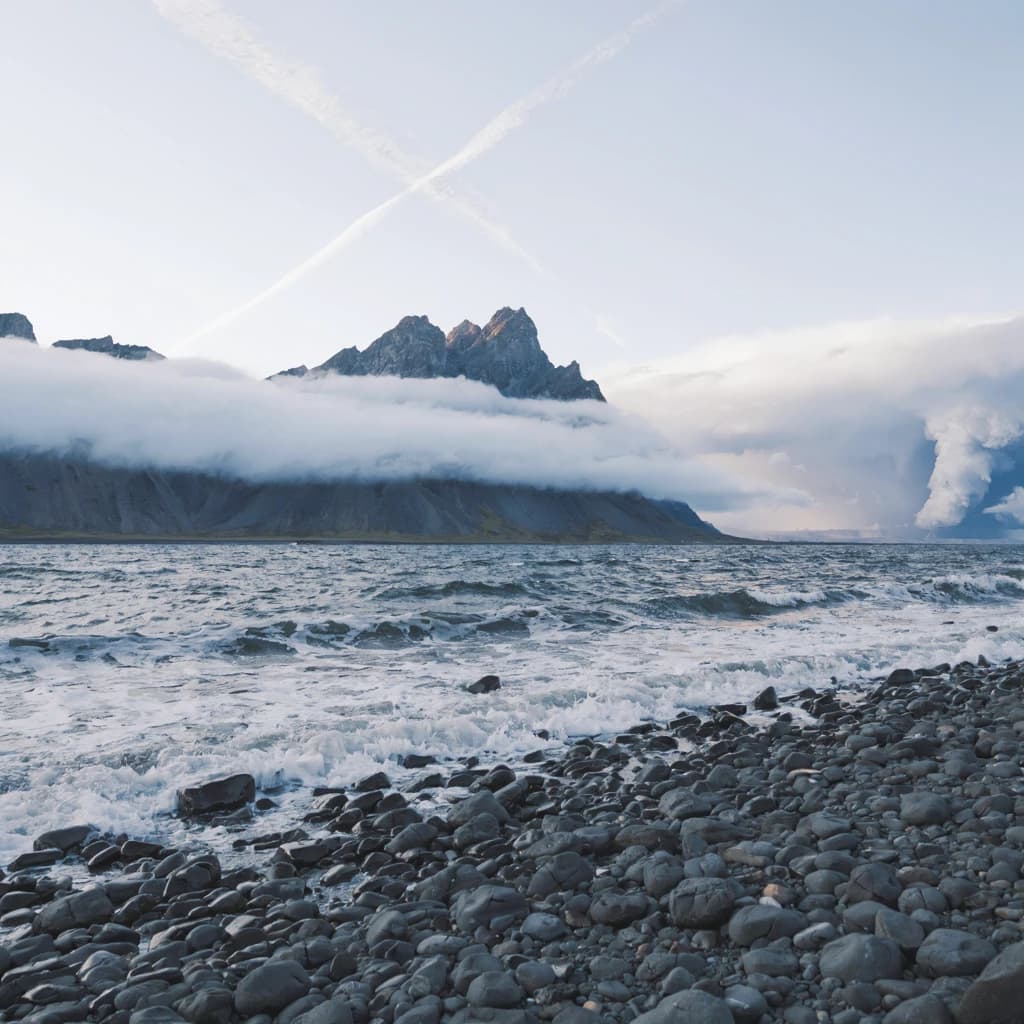 A stunning mountain vista pokes through the cloud top. Contrails from a distant airplane linger in the air. In the foreground there is a stony beach with foamy seas. A thunder storm is visibile in the distant right.