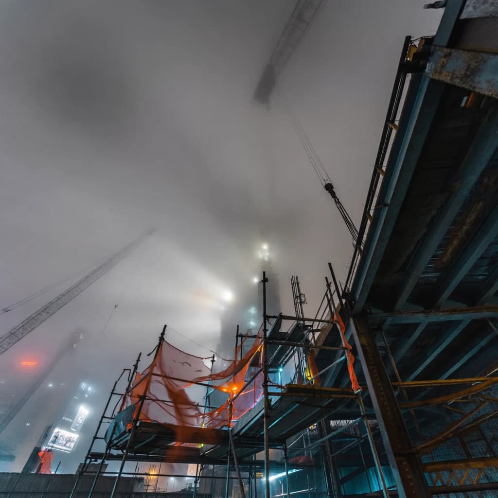 A construction site stretched upward into fog, cranes frozen in silhouette. Wind rattled scaffolding draped with torn mesh that glowed faintly from nearby billboards