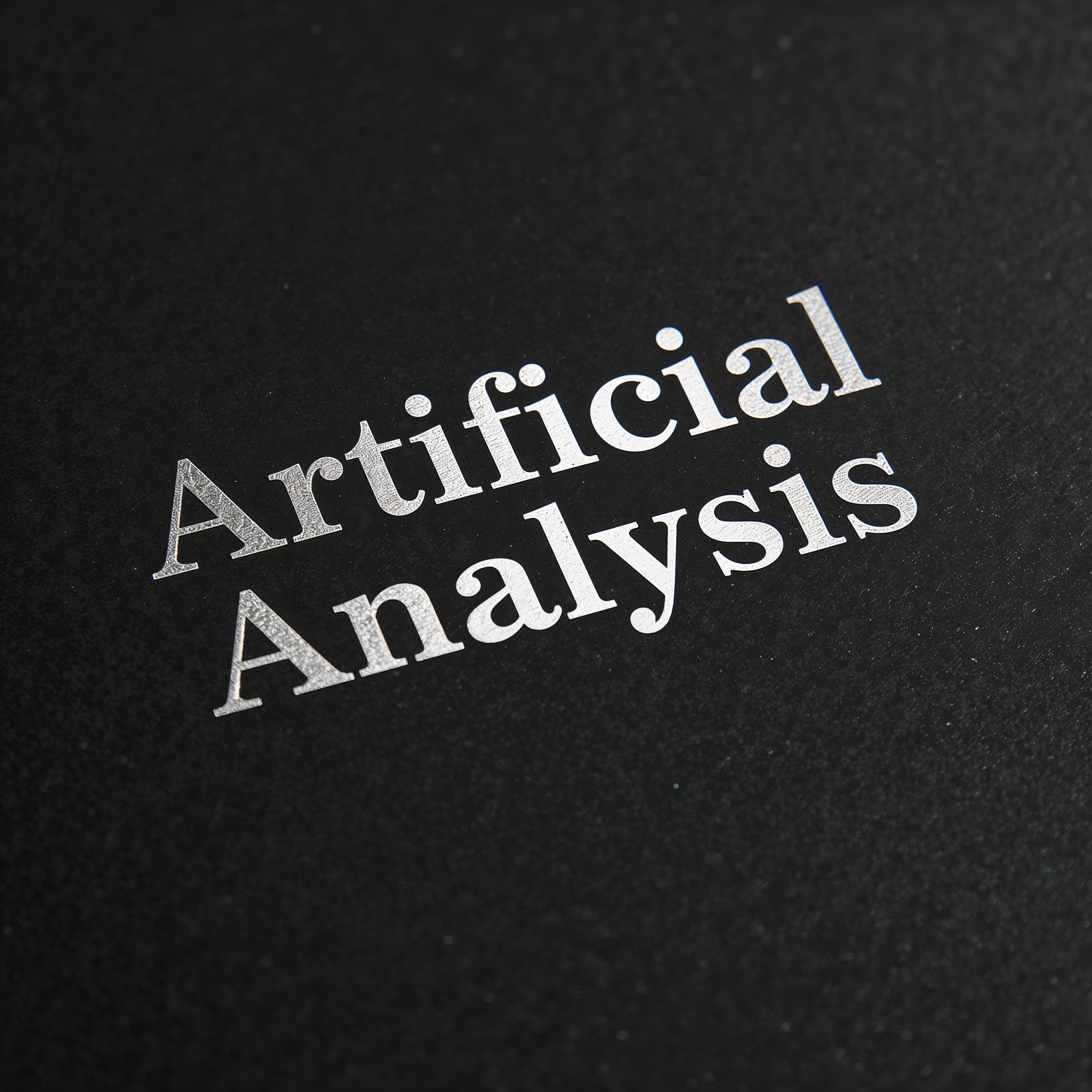 On the black box, silver foil embossing, centered: "Artificial Analysis" in Comic Sans Small Caps, perfectly justified. The letters are crisp, but the “Y” is duplicated faintly, ghosting right. On the box’s reflection, the word order is inverted: ARTIFICIAL ANALYSIS.