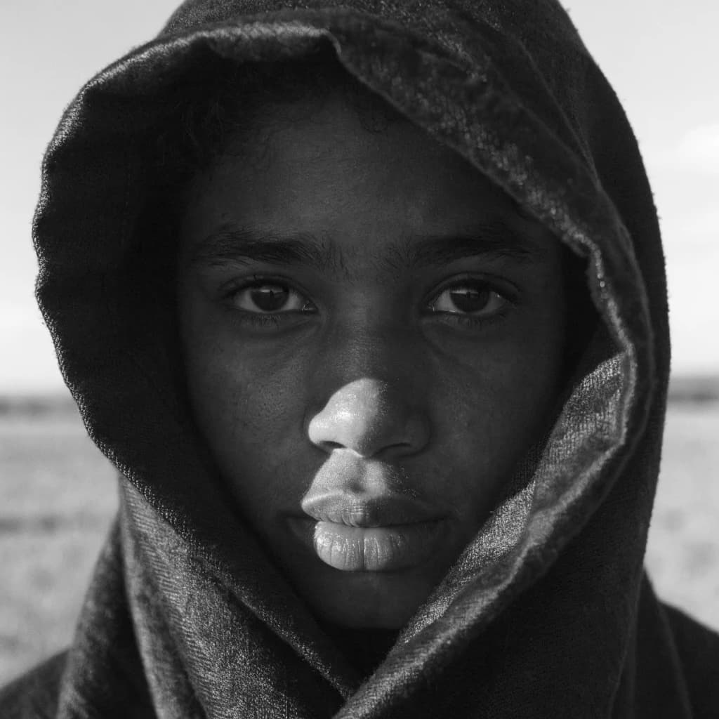 A dramatic black-and-white portrait of a young person draped in a hooded garment, their face framed by the hood's dark folds. Their skin tone and the subtle sheen on their cheeks create striking tonal contrast against the deep shadows. Their eyes are piercing, looking directly into the lens with a calm, steady intensity—neither confrontational nor submissive, but deeply present. The texture of the fabric is visible: it's dense, slightly weathered, and whisper-thin creases catch the light. In the background, an expansive, out-of-focus landscape suggests open space—perhaps distant plains or water—though the details remain soft and abstract. The overall mood is silent yet powerful, poised between vulnerability and resilience, with every highlight and shadow telling part of their story.