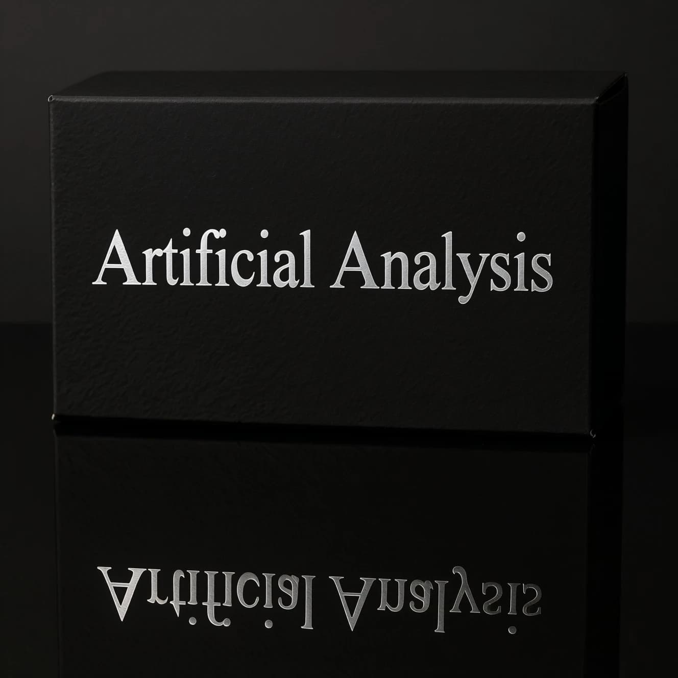 On the black box, silver foil embossing, centered: "Artificial Analysis" in Comic Sans Small Caps, perfectly justified. The letters are crisp, but the “Y” is duplicated faintly, ghosting right. On the box’s reflection, the word order is inverted: ARTIFICIAL ANALYSIS.