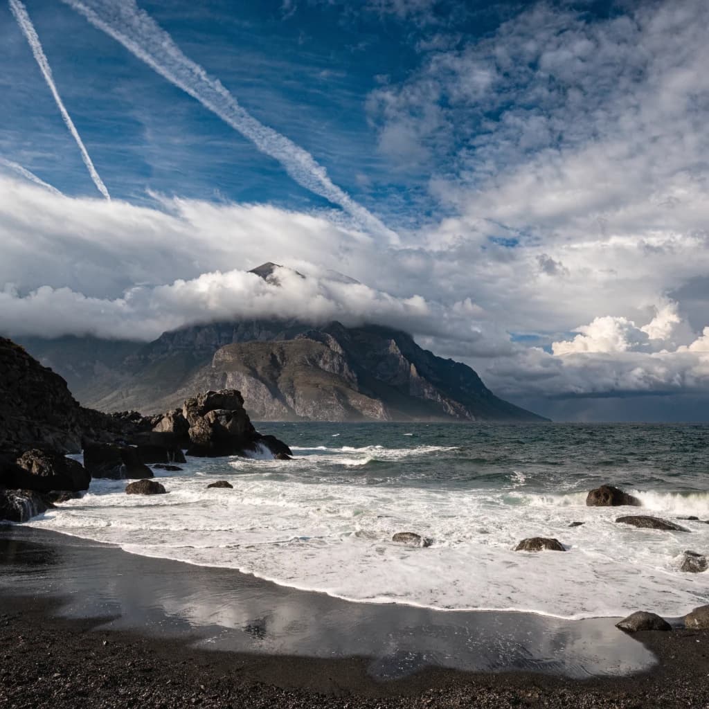 A stunning mountain vista pokes through the cloud top. Contrails from a distant airplane linger in the air. In the foreground there is a stony beach with foamy seas. A thunder storm is visibile in the distant right.