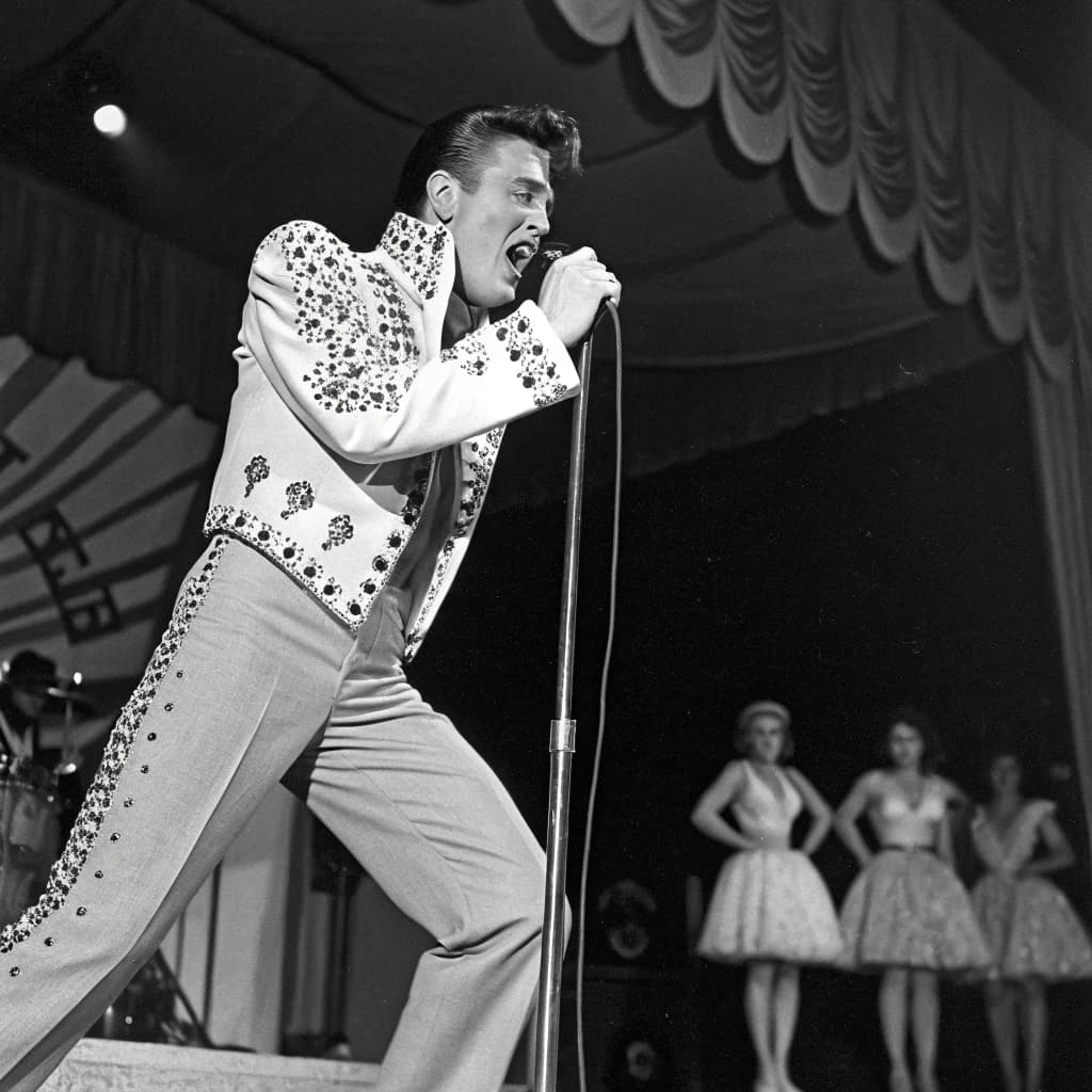 An archival image from 1953 of the rock singer swinging his hips on the stage while glamorous back up singers feign shock at his gyrations! He wears a diamond encrusted jacket with the stones arranged in the shape of a heart