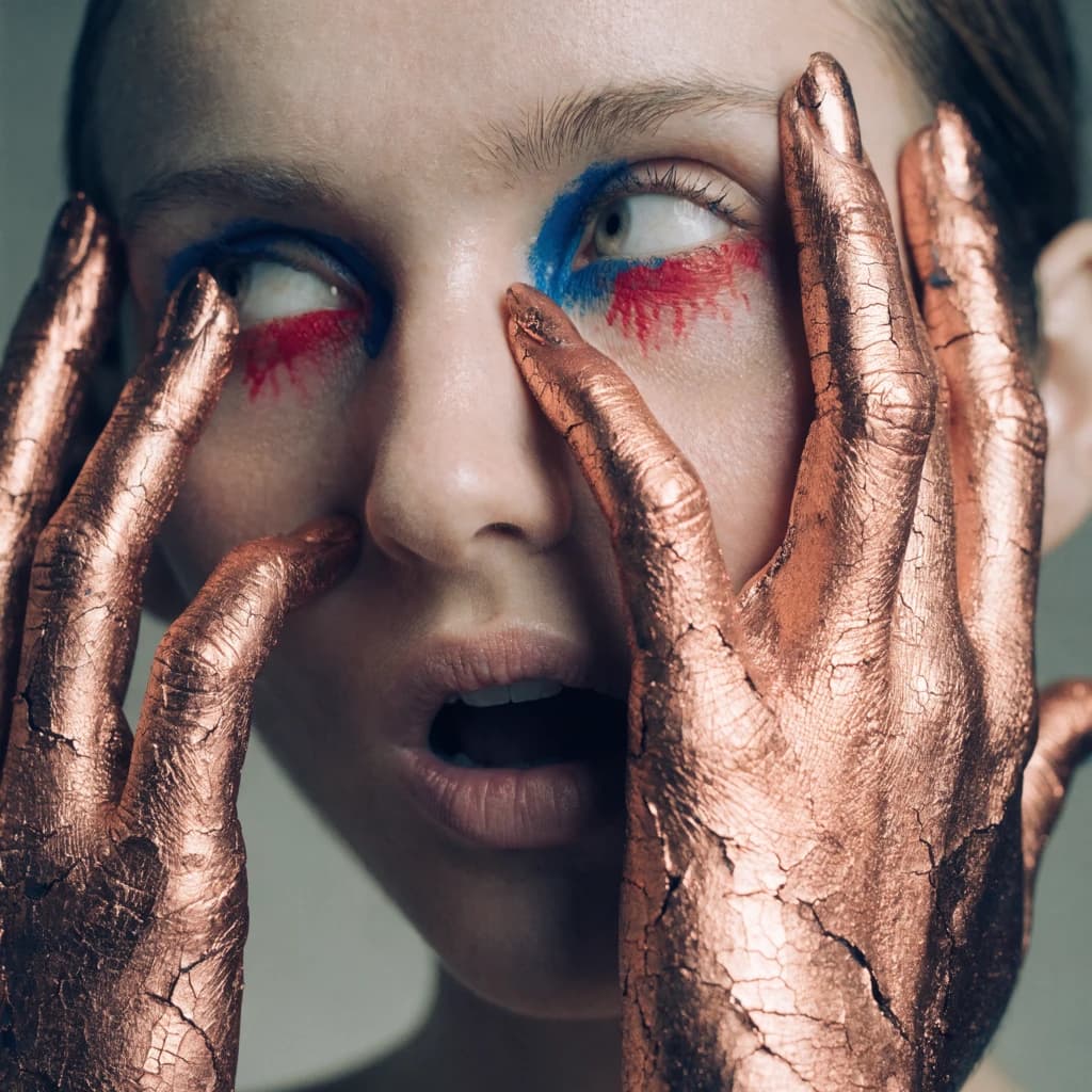 A striking close-up portrait of a woman with cracked metallic copper-painted hands framing her face. Her vivid blue and red eye makeup contrasts sharply with the smooth, pale skin and muted background, creating a bold, surreal composition. She displays an air of uncertainty about her