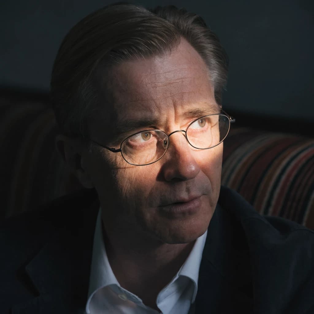 A sharply lit portrait of a middle-aged man wearing thin-rimmed glasses and a dark jacket over a white collared shirt. He gazes intently to the side, his face half illuminated by warm light while the background recedes into deep shadow. The expression is thoughtful, almost wary, as if caught mid-conversation. Behind him, a striped cushion in muted reds and blacks adds subtle texture, contrasting with the crisp highlights on his forehead and glasses. The overall mood is contemplative and cinematic, balancing warmth with tension.
