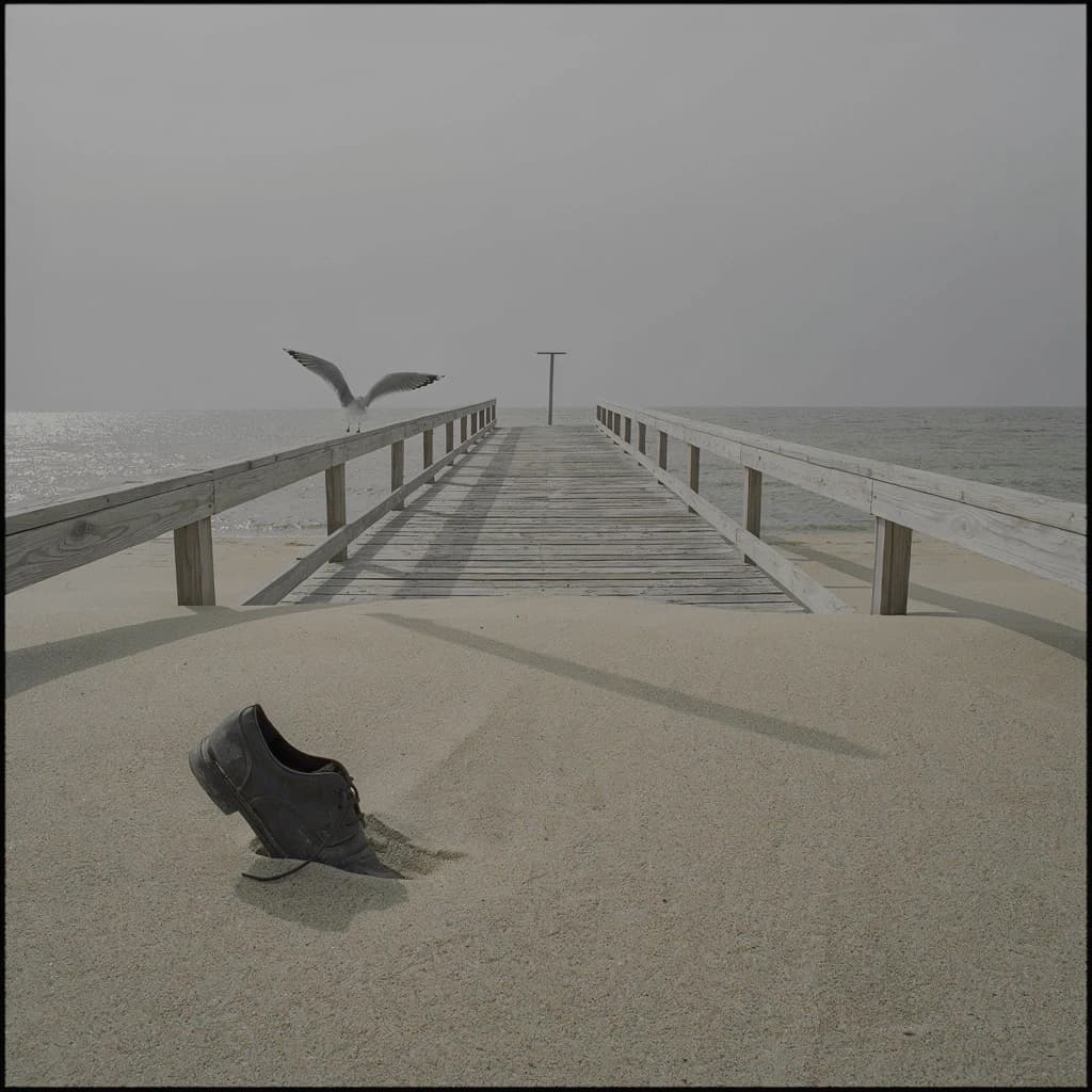 The beach is flat, sand beige, granular, no shells, except for a single left shoe, black leather, size eleven, half buried at an angle. The pier extends straight into the water, wood untreated, grain visible, though the support posts vanish before they touch the surface. The sky is uniformly gray, no clouds, yet shadows stretch at sharp diagonals. A single gull sits on the railing, wings outstretched, frozen mid-flap, no movement.