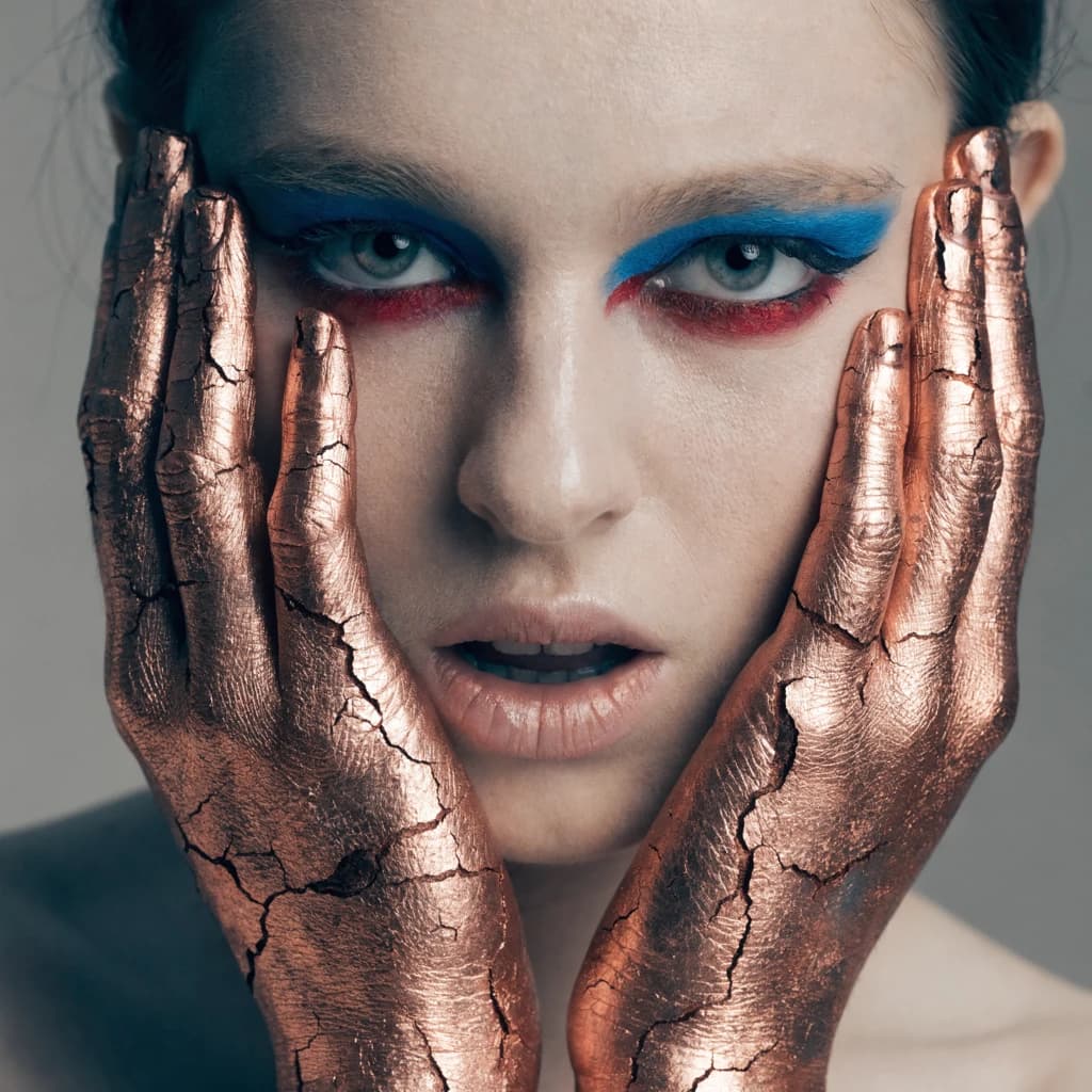 A striking close-up portrait of a woman with cracked metallic copper-painted hands framing her face. Her vivid blue and red eye makeup contrasts sharply with the smooth, pale skin and muted background, creating a bold, surreal composition. She displays an air of uncertainty about her