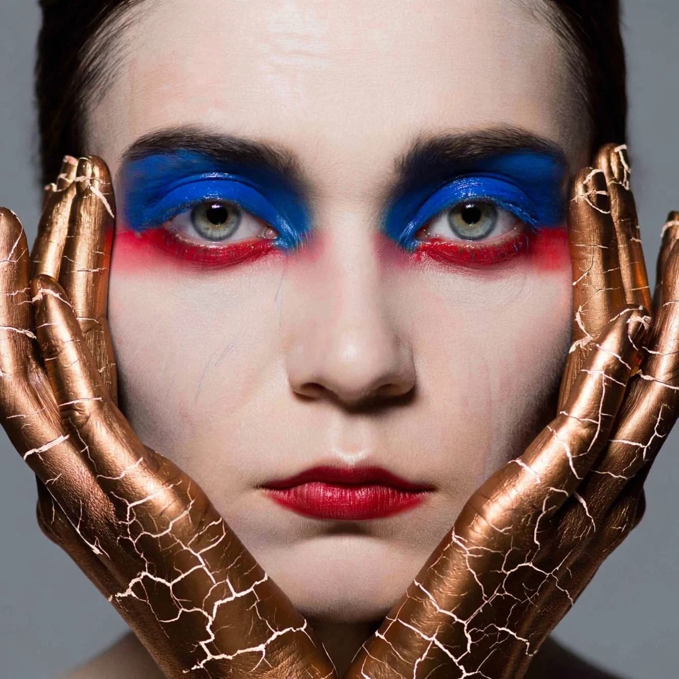 A striking close-up portrait of a woman with cracked metallic copper-painted hands framing her face. Her vivid blue and red eye makeup contrasts sharply with the smooth, pale skin and muted background, creating a bold, surreal composition. She displays an air of uncertainty about her