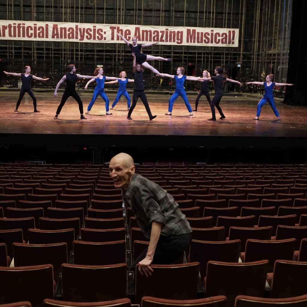 The musical troupe do a rehearsal in a large, empty theatre. 9 people are on the stage, half wear black and the others wear blue. The skinny bald director, seated in the second row, is practically leaping out of their seat with excitement as the leads nail their aerial. A giant banner reading "Artificial Analysis: The Amazing Musical!" is behind the actors.