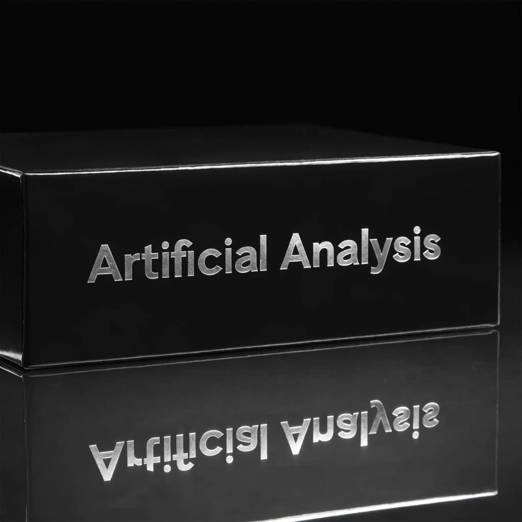 On the black box, silver foil embossing, centered: "Artificial Analysis" in Comic Sans Small Caps, perfectly justified. The letters are crisp, but the “Y” is duplicated faintly, ghosting right. On the box’s reflection, the word order is inverted: ARTIFICIAL ANALYSIS.