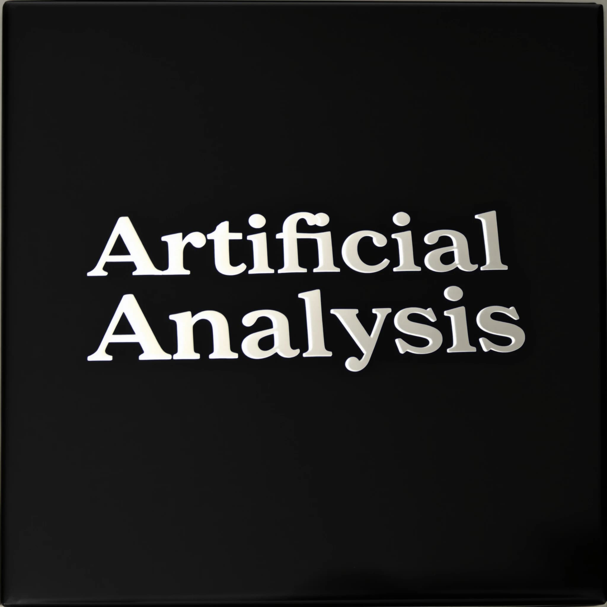 On the black box, silver foil embossing, centered: "Artificial Analysis" in Comic Sans Small Caps, perfectly justified. The letters are crisp, but the “Y” is duplicated faintly, ghosting right. On the box’s reflection, the word order is inverted: ARTIFICIAL ANALYSIS.
