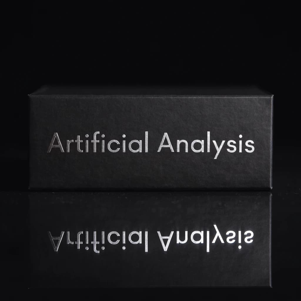 On the black box, silver foil embossing, centered: "Artificial Analysis" in Comic Sans Small Caps, perfectly justified. The letters are crisp, but the “Y” is duplicated faintly, ghosting right. On the box’s reflection, the word order is inverted: ARTIFICIAL ANALYSIS.