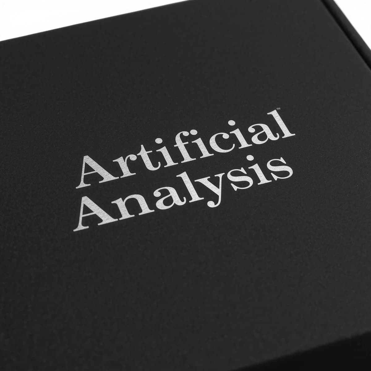 On the black box, silver foil embossing, centered: "Artificial Analysis" in Comic Sans Small Caps, perfectly justified. The letters are crisp, but the “Y” is duplicated faintly, ghosting right. On the box’s reflection, the word order is inverted: ARTIFICIAL ANALYSIS.