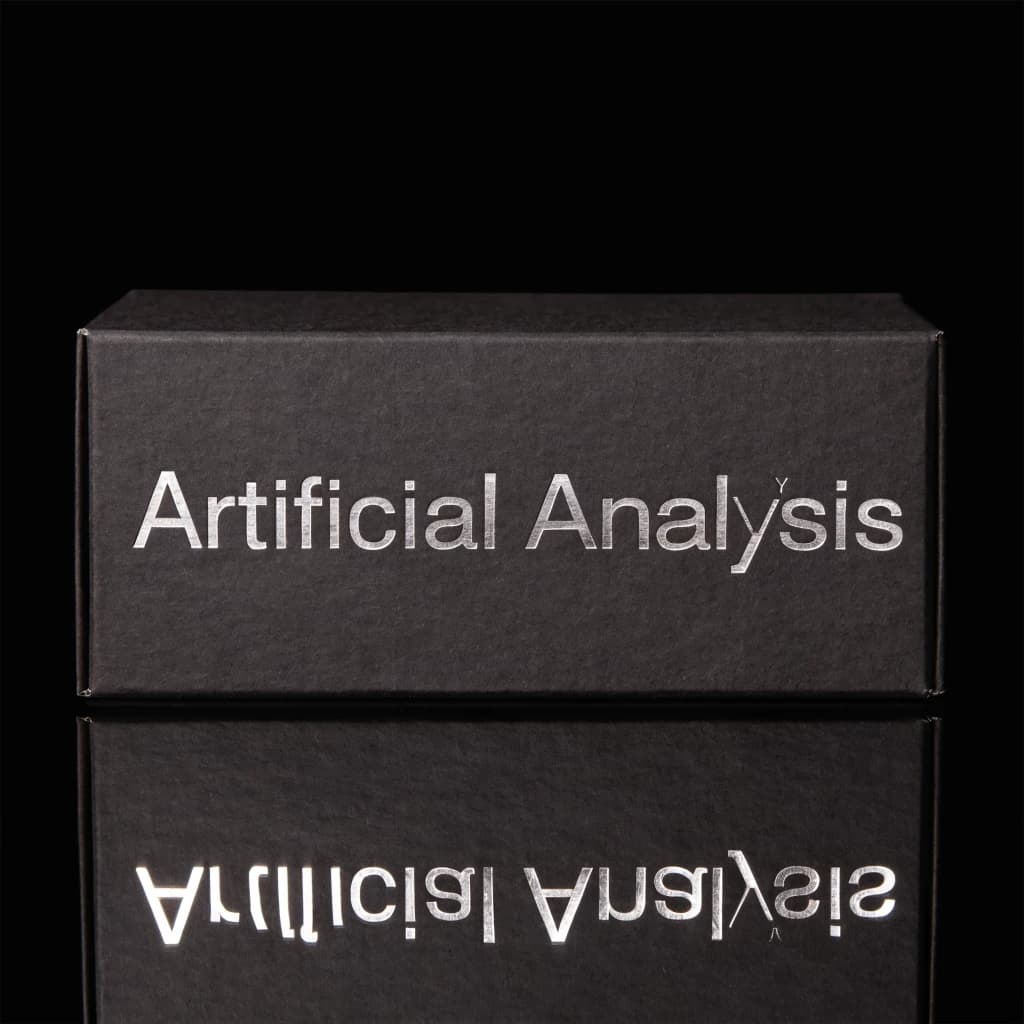 On the black box, silver foil embossing, centered: "Artificial Analysis" in Comic Sans Small Caps, perfectly justified. The letters are crisp, but the “Y” is duplicated faintly, ghosting right. On the box’s reflection, the word order is inverted: ARTIFICIAL ANALYSIS.