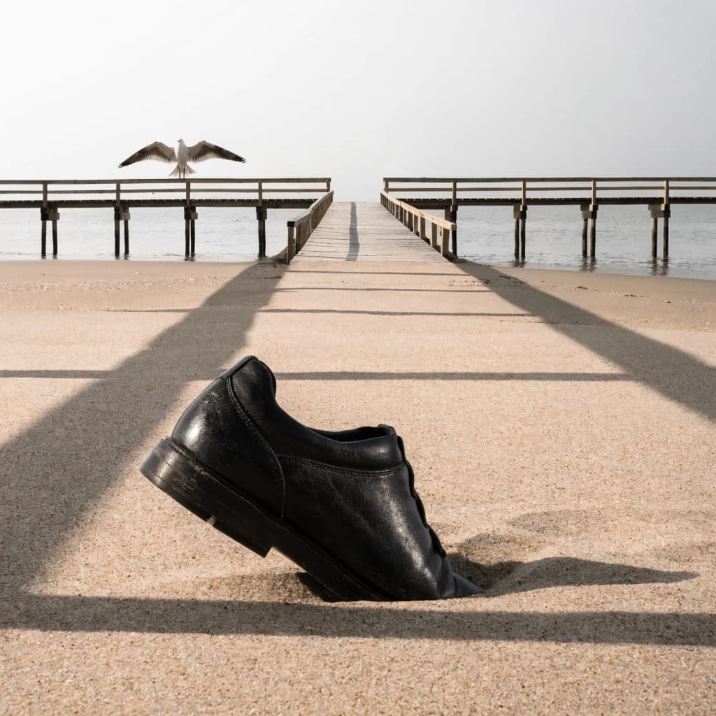 The beach is flat, sand beige, granular, no shells, except for a single left shoe, black leather, size eleven, half buried at an angle. The pier extends straight into the water, wood untreated, grain visible, though the support posts vanish before they touch the surface. The sky is uniformly gray, no clouds, yet shadows stretch at sharp diagonals. A single gull sits on the railing, wings outstretched, frozen mid-flap, no movement.