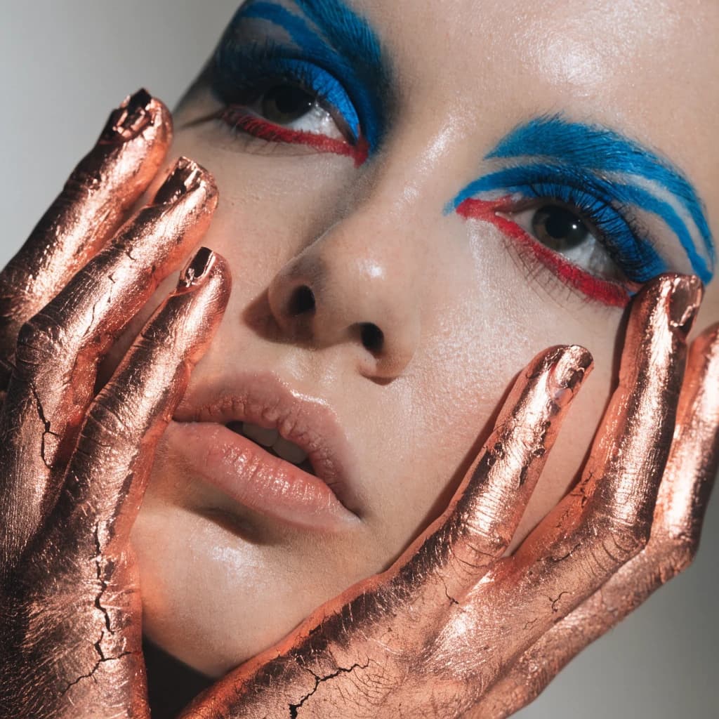 A striking close-up portrait of a woman with cracked metallic copper-painted hands framing her face. Her vivid blue and red eye makeup contrasts sharply with the smooth, pale skin and muted background, creating a bold, surreal composition. She displays an air of uncertainty about her