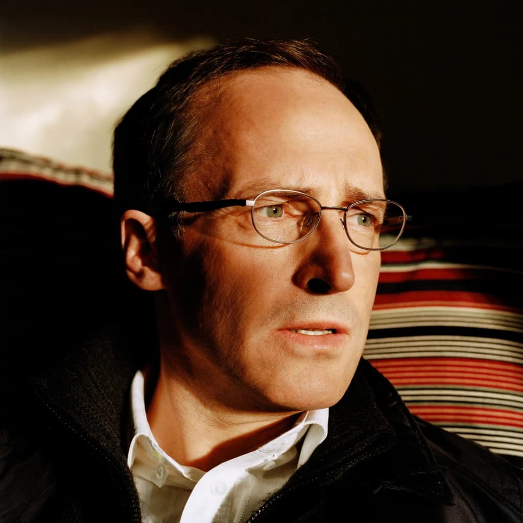 A sharply lit portrait of a middle-aged man wearing thin-rimmed glasses and a dark jacket over a white collared shirt. He gazes intently to the side, his face half illuminated by warm light while the background recedes into deep shadow. The expression is thoughtful, almost wary, as if caught mid-conversation. Behind him, a striped cushion in muted reds and blacks adds subtle texture, contrasting with the crisp highlights on his forehead and glasses. The overall mood is contemplative and cinematic, balancing warmth with tension.
