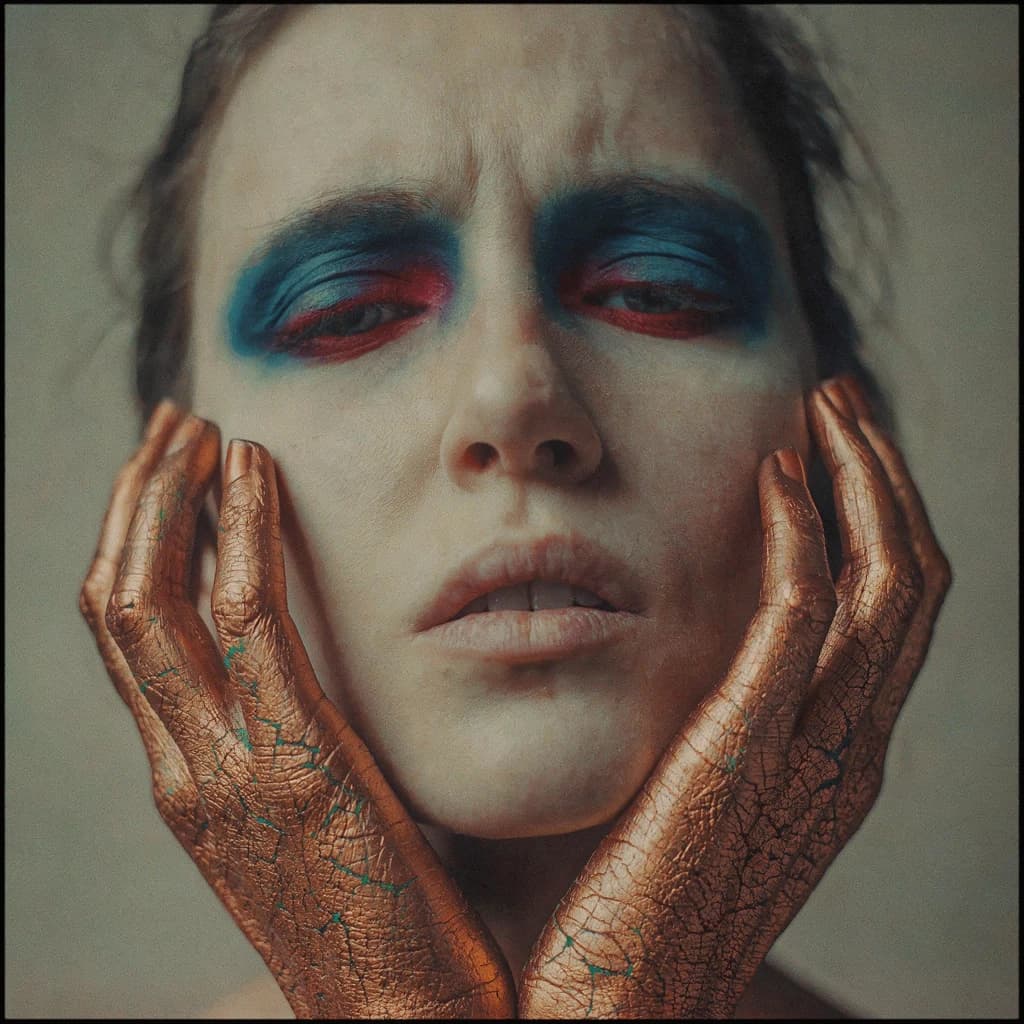 A striking close-up portrait of a woman with cracked metallic copper-painted hands framing her face. Her vivid blue and red eye makeup contrasts sharply with the smooth, pale skin and muted background, creating a bold, surreal composition. She displays an air of uncertainty about her
