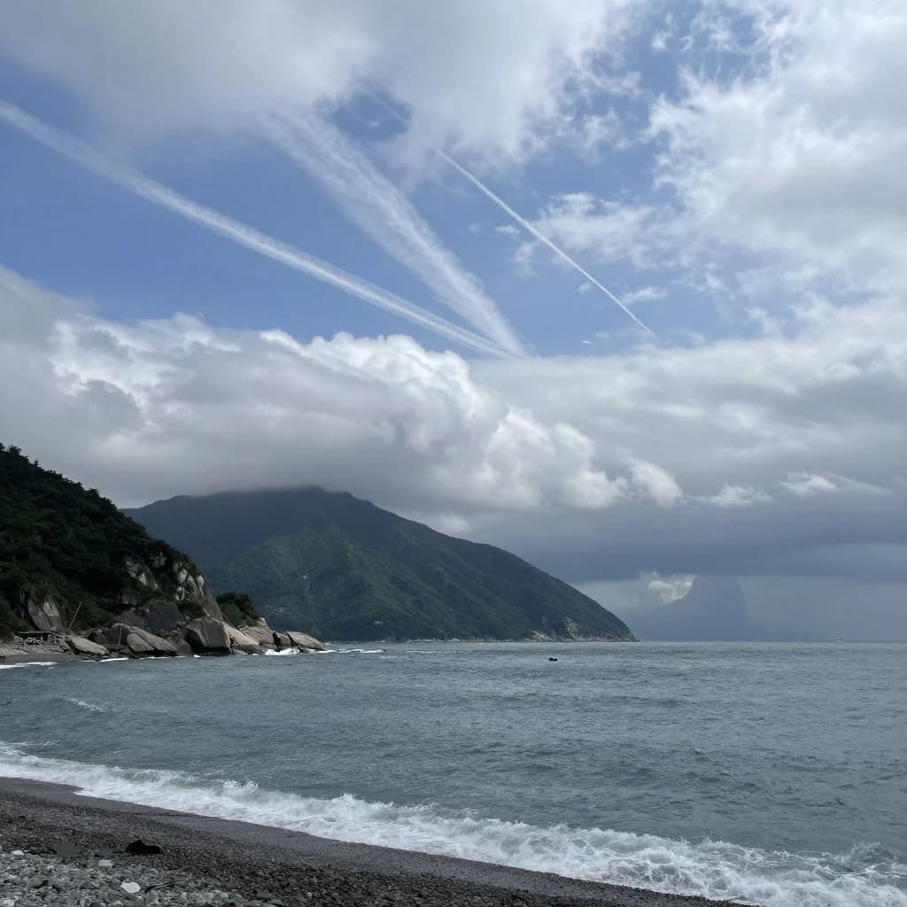 A stunning mountain vista pokes through the cloud top. Contrails from a distant airplane linger in the air. In the foreground there is a stony beach with foamy seas. A thunder storm is visibile in the distant right.