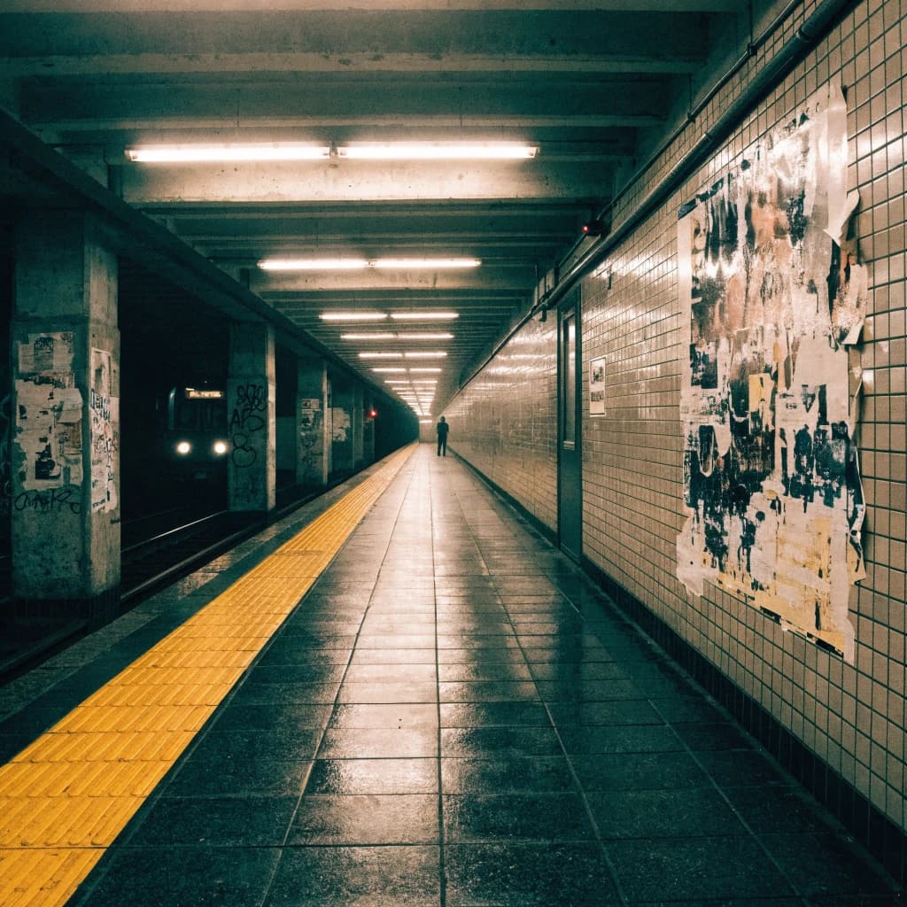 A subterranean subway platform with glossy tiles, peeling posters, flickering fluorescent lights, yellow safety line, and a distant train coming; slightly damp, echoes carry.