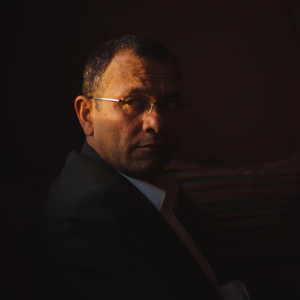 A sharply lit portrait of a middle-aged man wearing thin-rimmed glasses and a dark jacket over a white collared shirt. He gazes intently to the side, his face half illuminated by warm light while the background recedes into deep shadow. The expression is thoughtful, almost wary, as if caught mid-conversation. Behind him, a striped cushion in muted reds and blacks adds subtle texture, contrasting with the crisp highlights on his forehead and glasses. The overall mood is contemplative and cinematic, balancing warmth with tension.