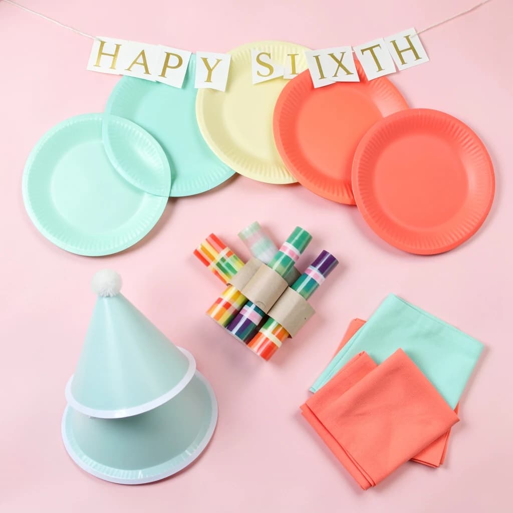 Flat lay photograph on pale pink paper, lit softly from above.
Eight paper plates fanned in an arc across the top of the frame, alternating mint and coral, four of each, mint first from the left.
Below the arc, twelve striped candles bundled under a paper band, laid diagonally.
Three cone party hats stacked at the lower left, only the middle hat trimmed with a pom pom.
At the lower right, six folded napkins in two piles of three, one pile mint and one coral.
A paper banner strung across the very top spells "HAPPY SIXTH" in gold letters, a few letters overlapping the plate arc.