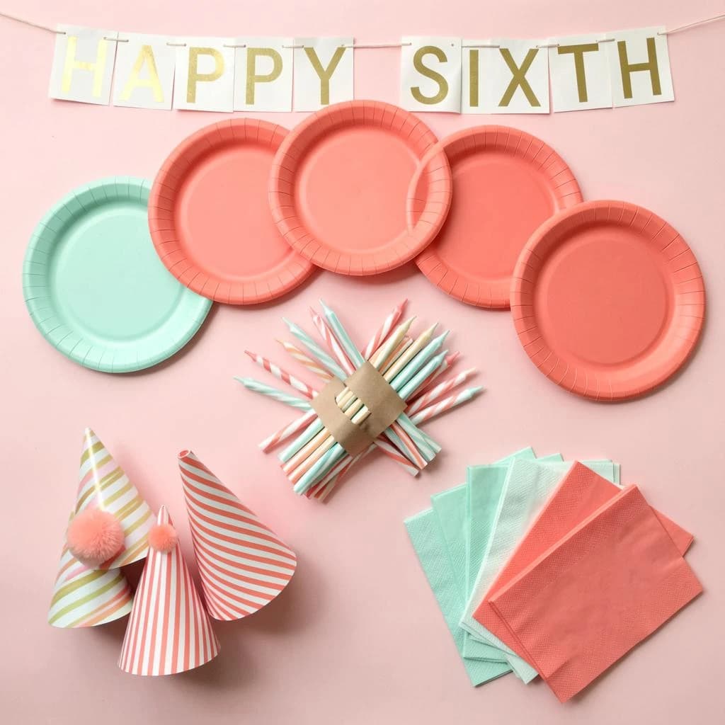 Flat lay photograph on pale pink paper, lit softly from above.
Eight paper plates fanned in an arc across the top of the frame, alternating mint and coral, four of each, mint first from the left.
Below the arc, twelve striped candles bundled under a paper band, laid diagonally.
Three cone party hats stacked at the lower left, only the middle hat trimmed with a pom pom.
At the lower right, six folded napkins in two piles of three, one pile mint and one coral.
A paper banner strung across the very top spells "HAPPY SIXTH" in gold letters, a few letters overlapping the plate arc.