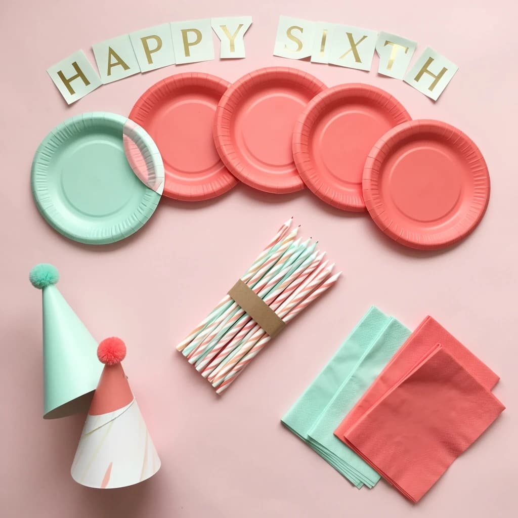Flat lay photograph on pale pink paper, lit softly from above.
Eight paper plates fanned in an arc across the top of the frame, alternating mint and coral, four of each, mint first from the left.
Below the arc, twelve striped candles bundled under a paper band, laid diagonally.
Three cone party hats stacked at the lower left, only the middle hat trimmed with a pom pom.
At the lower right, six folded napkins in two piles of three, one pile mint and one coral.
A paper banner strung across the very top spells "HAPPY SIXTH" in gold letters, a few letters overlapping the plate arc.