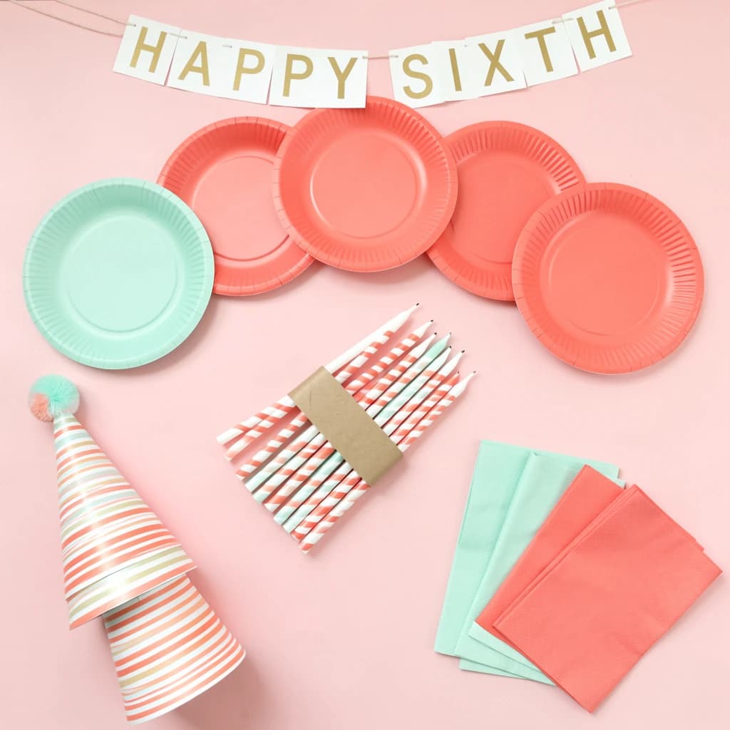 Flat lay photograph on pale pink paper, lit softly from above.
Eight paper plates fanned in an arc across the top of the frame, alternating mint and coral, four of each, mint first from the left.
Below the arc, twelve striped candles bundled under a paper band, laid diagonally.
Three cone party hats stacked at the lower left, only the middle hat trimmed with a pom pom.
At the lower right, six folded napkins in two piles of three, one pile mint and one coral.
A paper banner strung across the very top spells "HAPPY SIXTH" in gold letters, a few letters overlapping the plate arc.