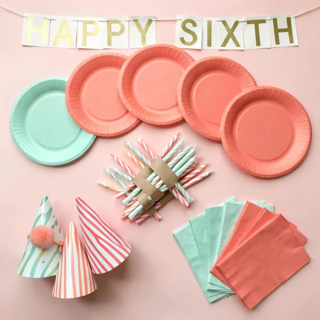 Flat lay photograph on pale pink paper, lit softly from above.
Eight paper plates fanned in an arc across the top of the frame, alternating mint and coral, four of each, mint first from the left.
Below the arc, twelve striped candles bundled under a paper band, laid diagonally.
Three cone party hats stacked at the lower left, only the middle hat trimmed with a pom pom.
At the lower right, six folded napkins in two piles of three, one pile mint and one coral.
A paper banner strung across the very top spells "HAPPY SIXTH" in gold letters, a few letters overlapping the plate arc.