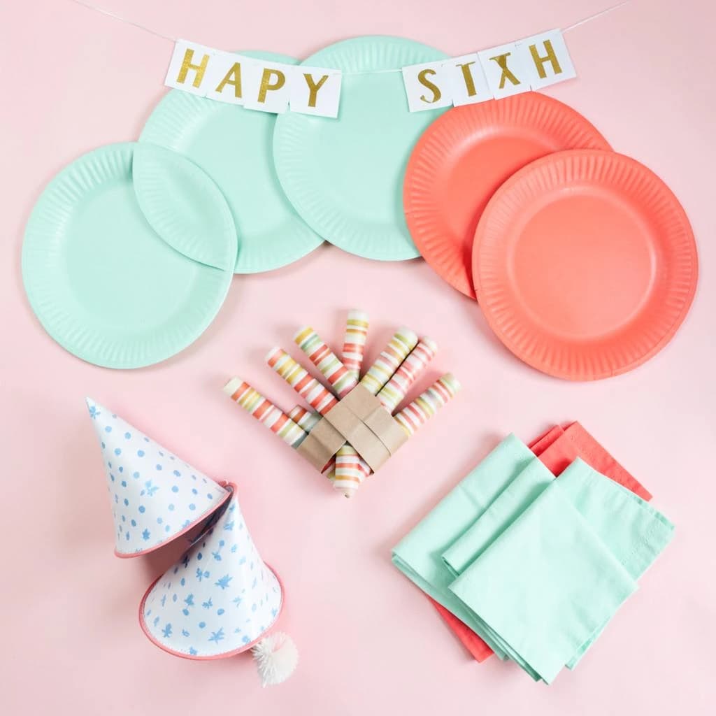 Flat lay photograph on pale pink paper, lit softly from above.
Eight paper plates fanned in an arc across the top of the frame, alternating mint and coral, four of each, mint first from the left.
Below the arc, twelve striped candles bundled under a paper band, laid diagonally.
Three cone party hats stacked at the lower left, only the middle hat trimmed with a pom pom.
At the lower right, six folded napkins in two piles of three, one pile mint and one coral.
A paper banner strung across the very top spells "HAPPY SIXTH" in gold letters, a few letters overlapping the plate arc.