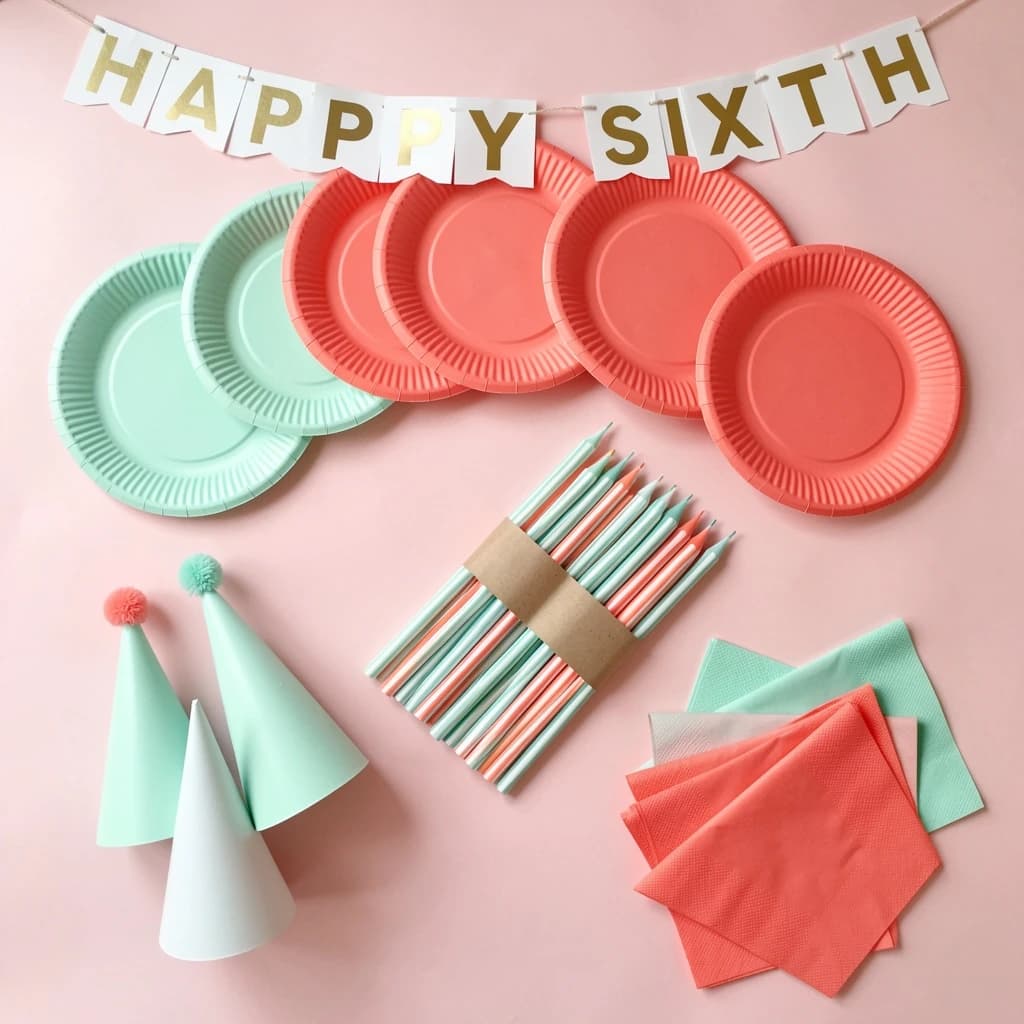 Flat lay photograph on pale pink paper, lit softly from above.
Eight paper plates fanned in an arc across the top of the frame, alternating mint and coral, four of each, mint first from the left.
Below the arc, twelve striped candles bundled under a paper band, laid diagonally.
Three cone party hats stacked at the lower left, only the middle hat trimmed with a pom pom.
At the lower right, six folded napkins in two piles of three, one pile mint and one coral.
A paper banner strung across the very top spells "HAPPY SIXTH" in gold letters, a few letters overlapping the plate arc.