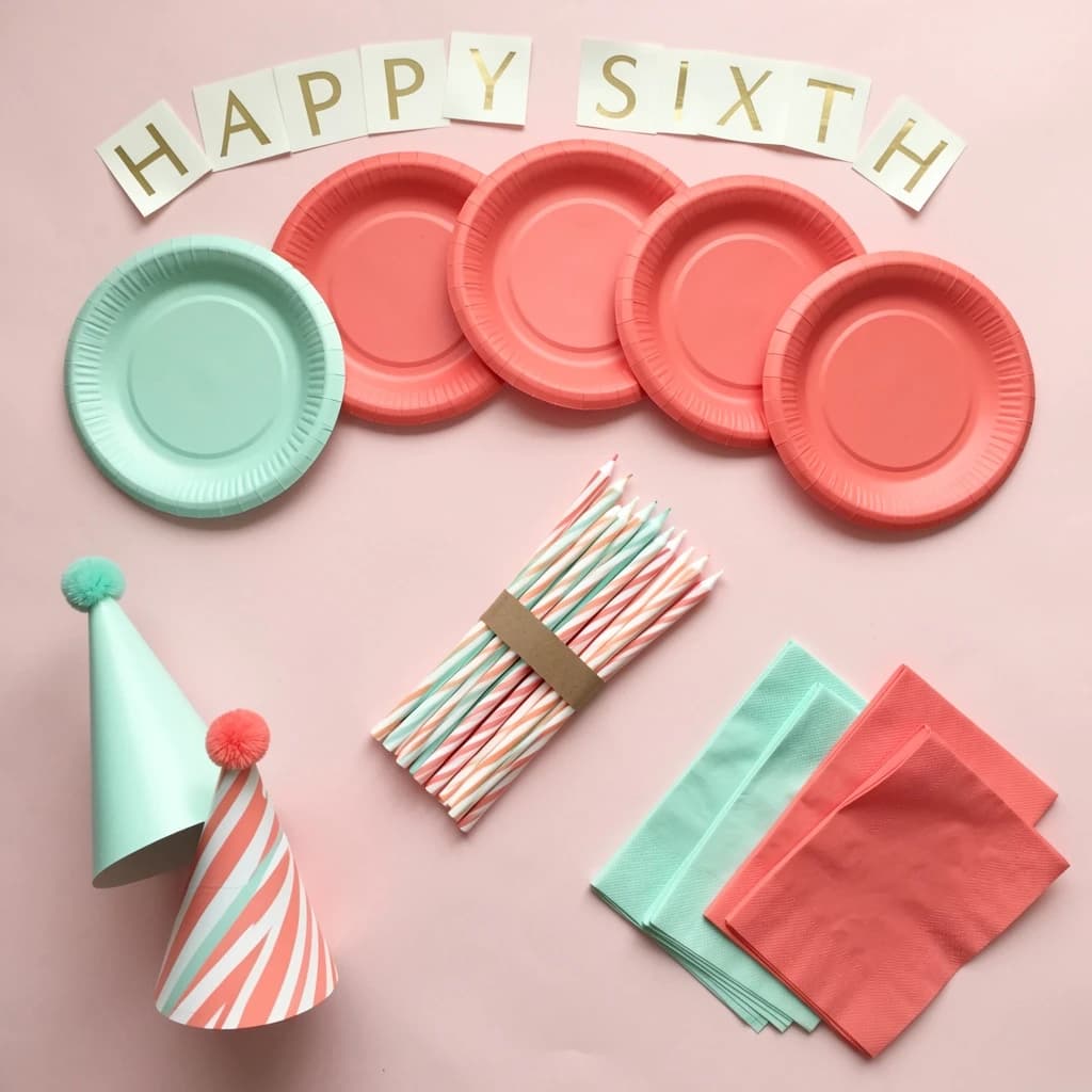 Flat lay photograph on pale pink paper, lit softly from above.
Eight paper plates fanned in an arc across the top of the frame, alternating mint and coral, four of each, mint first from the left.
Below the arc, twelve striped candles bundled under a paper band, laid diagonally.
Three cone party hats stacked at the lower left, only the middle hat trimmed with a pom pom.
At the lower right, six folded napkins in two piles of three, one pile mint and one coral.
A paper banner strung across the very top spells "HAPPY SIXTH" in gold letters, a few letters overlapping the plate arc.
