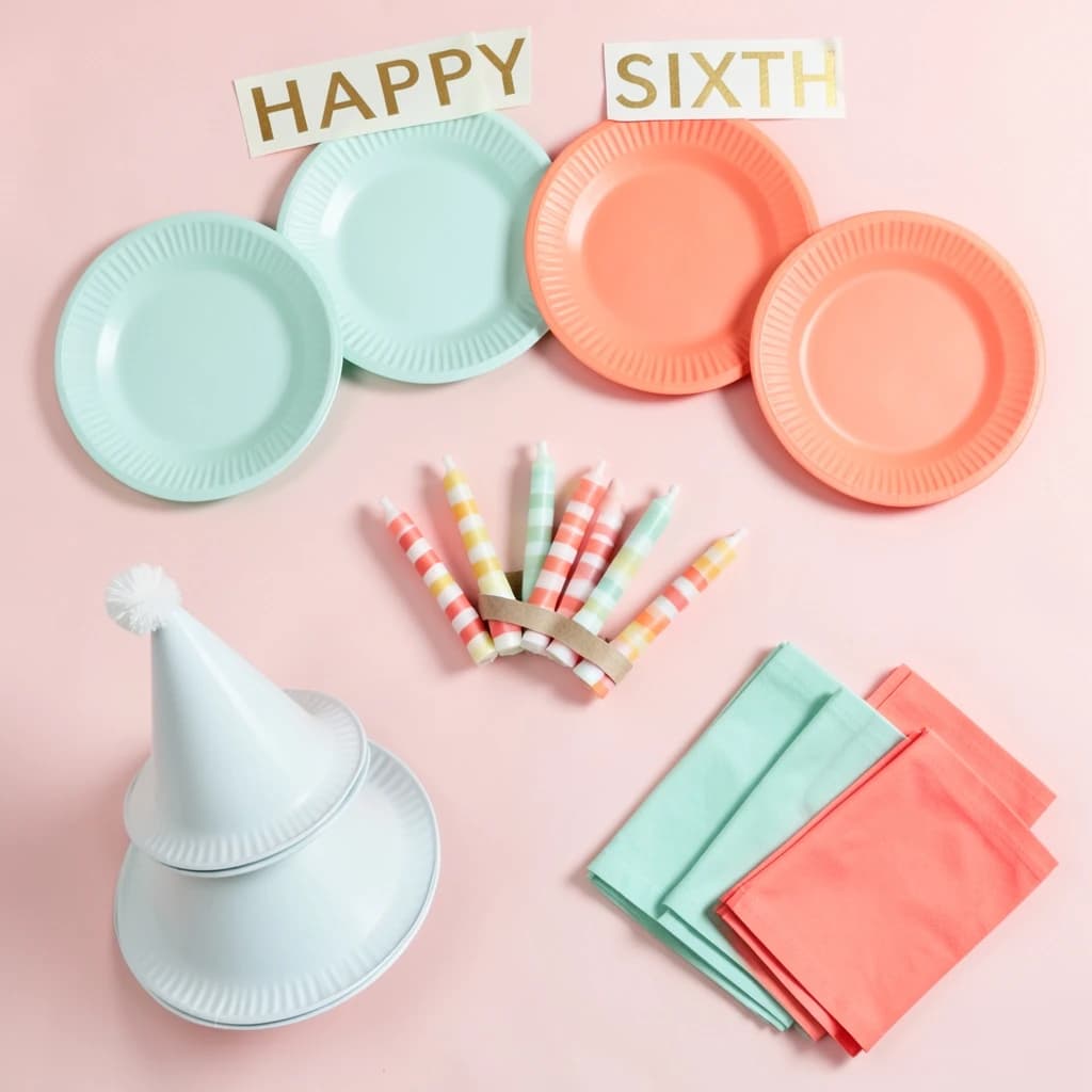 Flat lay photograph on pale pink paper, lit softly from above.
Eight paper plates fanned in an arc across the top of the frame, alternating mint and coral, four of each, mint first from the left.
Below the arc, twelve striped candles bundled under a paper band, laid diagonally.
Three cone party hats stacked at the lower left, only the middle hat trimmed with a pom pom.
At the lower right, six folded napkins in two piles of three, one pile mint and one coral.
A paper banner strung across the very top spells "HAPPY SIXTH" in gold letters, a few letters overlapping the plate arc.
