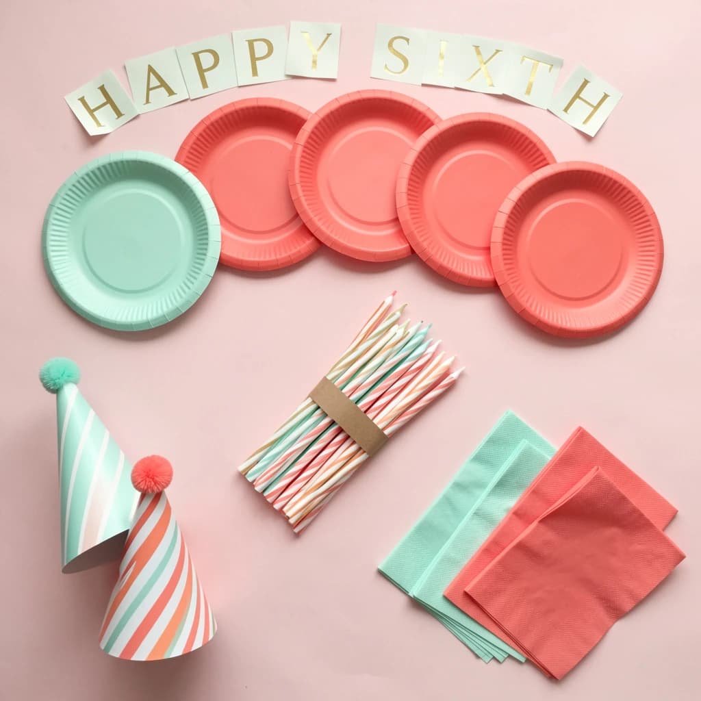 Flat lay photograph on pale pink paper, lit softly from above.
Eight paper plates fanned in an arc across the top of the frame, alternating mint and coral, four of each, mint first from the left.
Below the arc, twelve striped candles bundled under a paper band, laid diagonally.
Three cone party hats stacked at the lower left, only the middle hat trimmed with a pom pom.
At the lower right, six folded napkins in two piles of three, one pile mint and one coral.
A paper banner strung across the very top spells "HAPPY SIXTH" in gold letters, a few letters overlapping the plate arc.