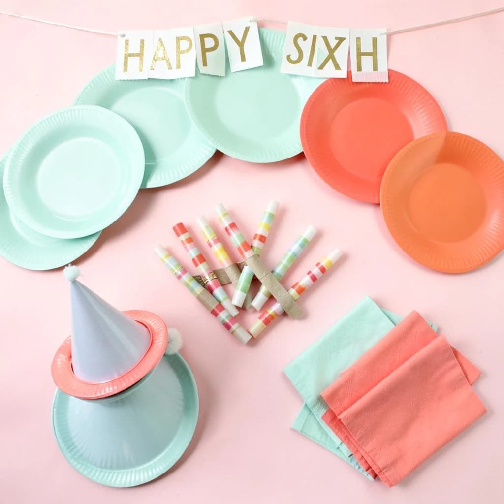 Flat lay photograph on pale pink paper, lit softly from above.
Eight paper plates fanned in an arc across the top of the frame, alternating mint and coral, four of each, mint first from the left.
Below the arc, twelve striped candles bundled under a paper band, laid diagonally.
Three cone party hats stacked at the lower left, only the middle hat trimmed with a pom pom.
At the lower right, six folded napkins in two piles of three, one pile mint and one coral.
A paper banner strung across the very top spells "HAPPY SIXTH" in gold letters, a few letters overlapping the plate arc.