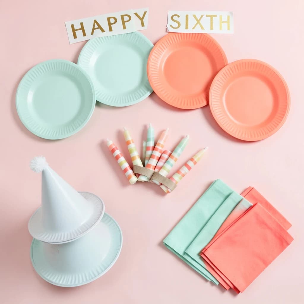 Flat lay photograph on pale pink paper, lit softly from above.
Eight paper plates fanned in an arc across the top of the frame, alternating mint and coral, four of each, mint first from the left.
Below the arc, twelve striped candles bundled under a paper band, laid diagonally.
Three cone party hats stacked at the lower left, only the middle hat trimmed with a pom pom.
At the lower right, six folded napkins in two piles of three, one pile mint and one coral.
A paper banner strung across the very top spells "HAPPY SIXTH" in gold letters, a few letters overlapping the plate arc.