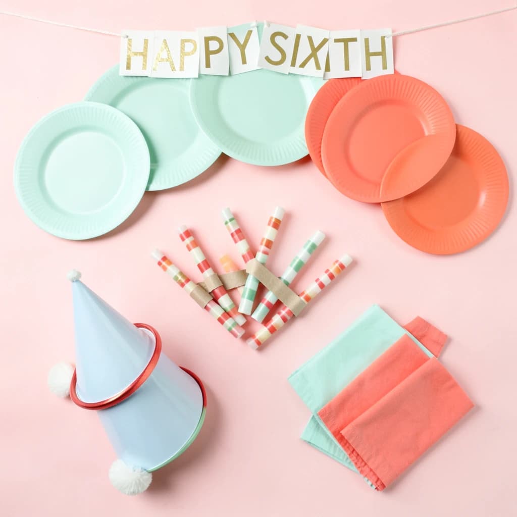 Flat lay photograph on pale pink paper, lit softly from above.
Eight paper plates fanned in an arc across the top of the frame, alternating mint and coral, four of each, mint first from the left.
Below the arc, twelve striped candles bundled under a paper band, laid diagonally.
Three cone party hats stacked at the lower left, only the middle hat trimmed with a pom pom.
At the lower right, six folded napkins in two piles of three, one pile mint and one coral.
A paper banner strung across the very top spells "HAPPY SIXTH" in gold letters, a few letters overlapping the plate arc.
