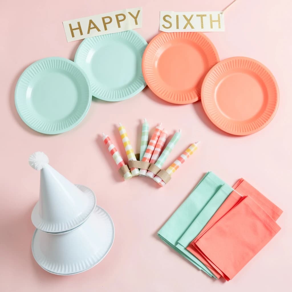 Flat lay photograph on pale pink paper, lit softly from above.
Eight paper plates fanned in an arc across the top of the frame, alternating mint and coral, four of each, mint first from the left.
Below the arc, twelve striped candles bundled under a paper band, laid diagonally.
Three cone party hats stacked at the lower left, only the middle hat trimmed with a pom pom.
At the lower right, six folded napkins in two piles of three, one pile mint and one coral.
A paper banner strung across the very top spells "HAPPY SIXTH" in gold letters, a few letters overlapping the plate arc.