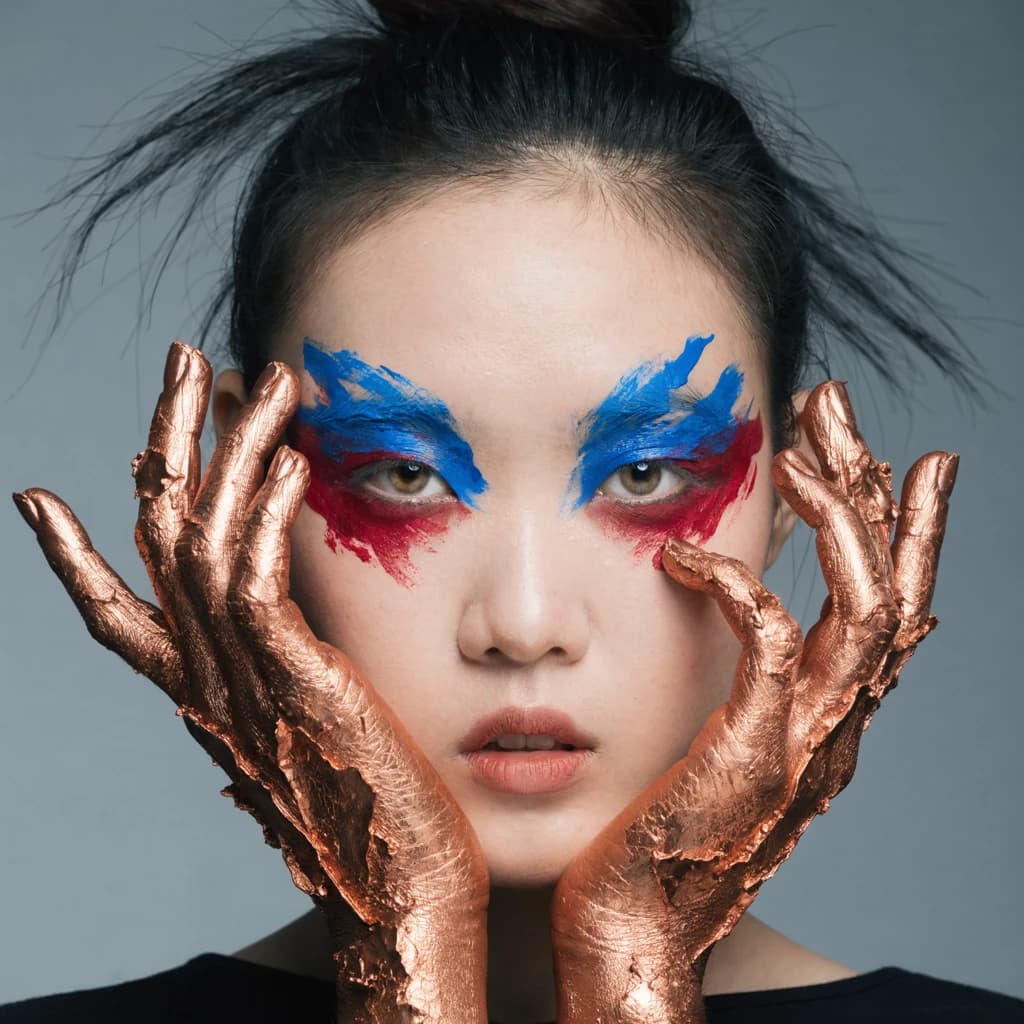A striking close-up portrait of a woman with cracked metallic copper-painted hands framing her face. Her vivid blue and red eye makeup contrasts sharply with the smooth, pale skin and muted background, creating a bold, surreal composition. She displays an air of uncertainty about her