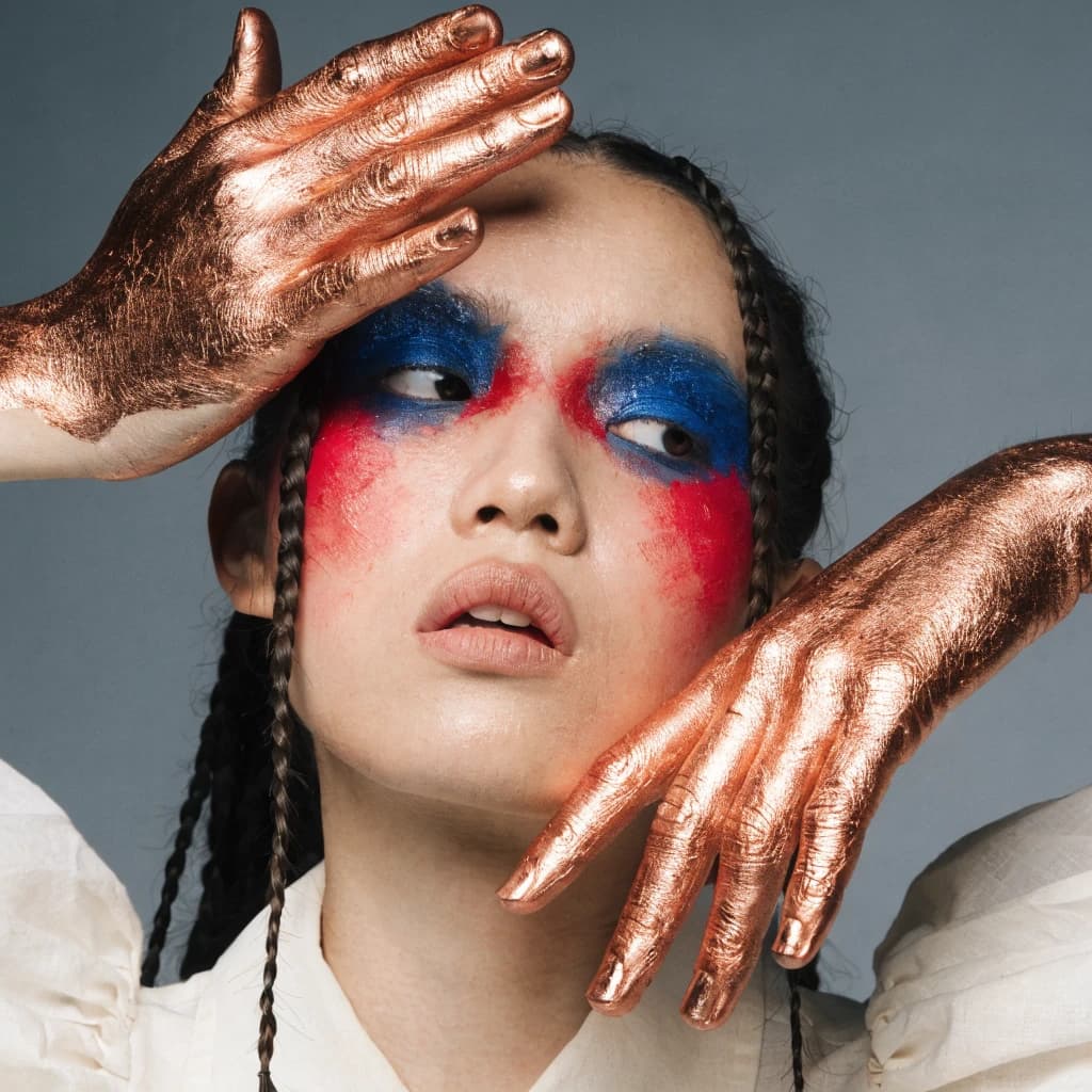 A striking close-up portrait of a woman with cracked metallic copper-painted hands framing her face. Her vivid blue and red eye makeup contrasts sharply with the smooth, pale skin and muted background, creating a bold, surreal composition. She displays an air of uncertainty about her