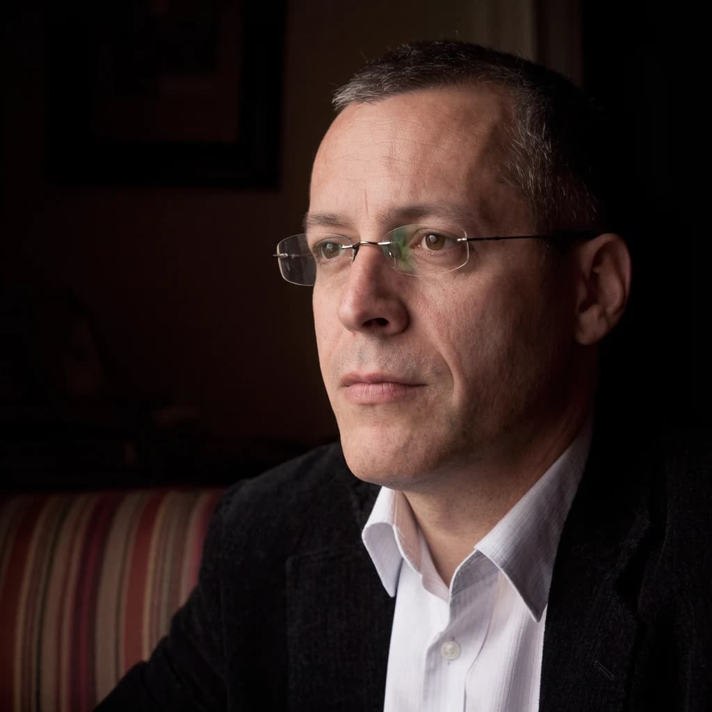 A sharply lit portrait of a middle-aged man wearing thin-rimmed glasses and a dark jacket over a white collared shirt. He gazes intently to the side, his face half illuminated by warm light while the background recedes into deep shadow. The expression is thoughtful, almost wary, as if caught mid-conversation. Behind him, a striped cushion in muted reds and blacks adds subtle texture, contrasting with the crisp highlights on his forehead and glasses. The overall mood is contemplative and cinematic, balancing warmth with tension.