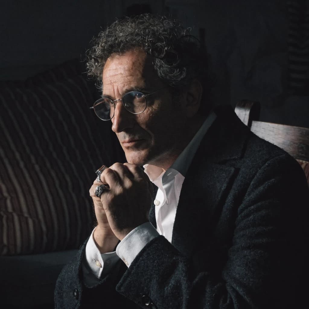 A sharply lit portrait of a middle-aged man wearing thin-rimmed glasses and a dark jacket over a white collared shirt. He gazes intently to the side, his face half illuminated by warm light while the background recedes into deep shadow. The expression is thoughtful, almost wary, as if caught mid-conversation. Behind him, a striped cushion in muted reds and blacks adds subtle texture, contrasting with the crisp highlights on his forehead and glasses. The overall mood is contemplative and cinematic, balancing warmth with tension.